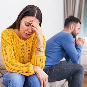 An upset woman sits on a couch, holding her head, while a man sits behind her looking away, Represented Divorce Cases.